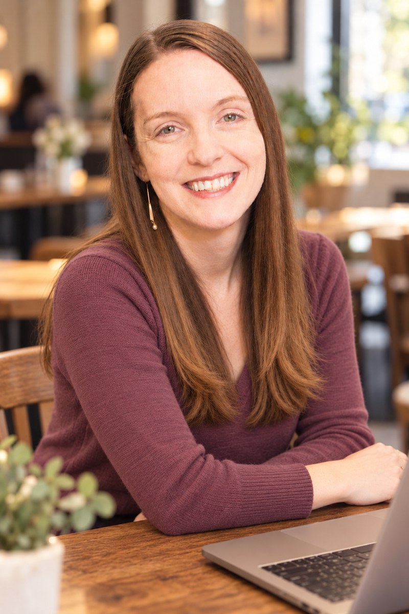 Portrait of Ashley, smiling, seated at a café table in a plum sweater